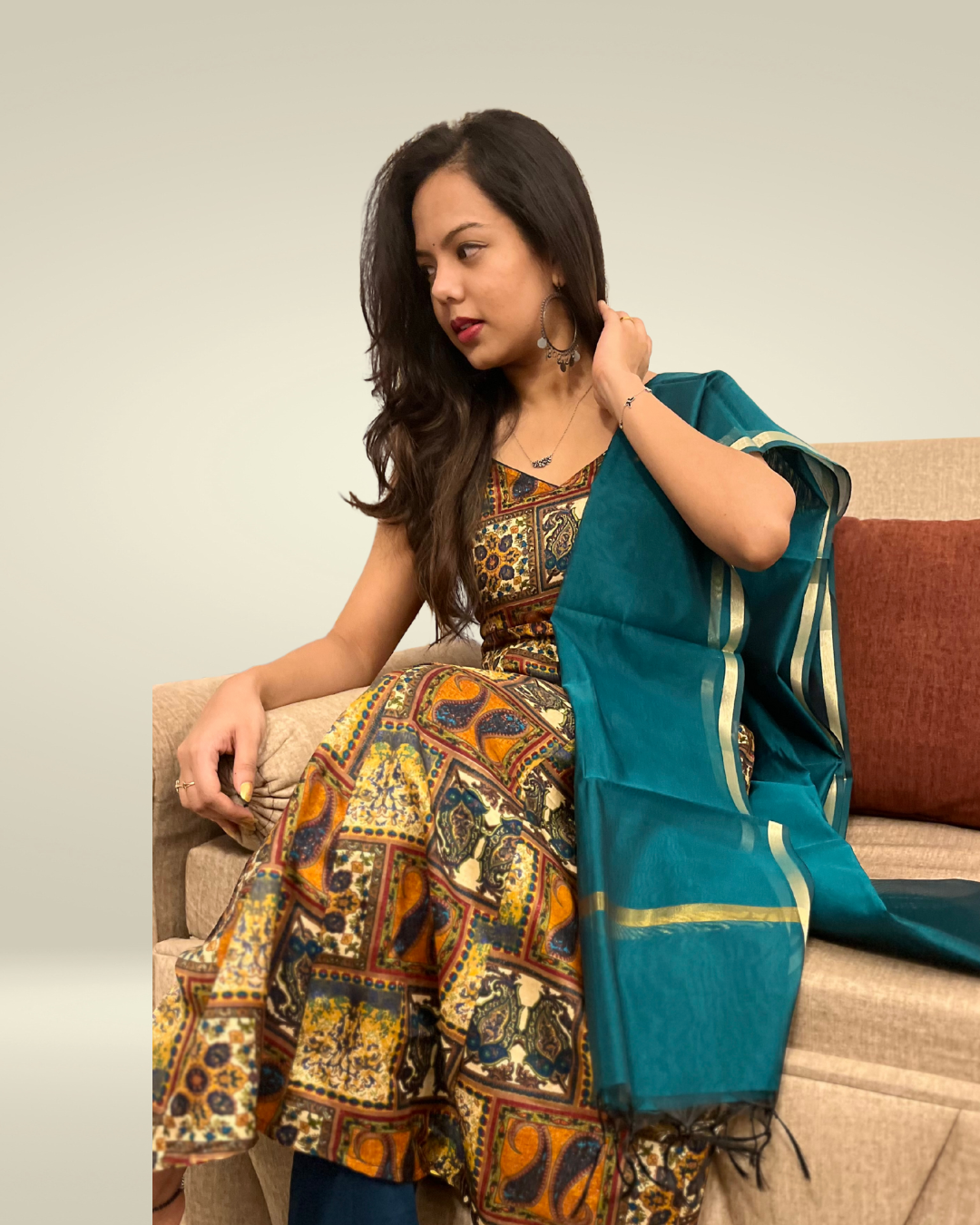 Woman sitting on a couch wearing a patterned dress and teal saree.